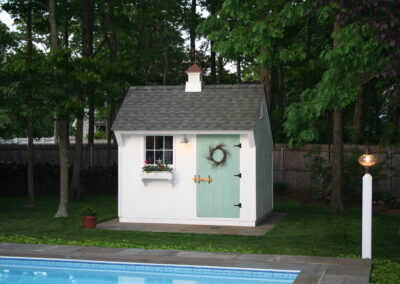 White Pool Shed with green door with lamp post and pool in the background
